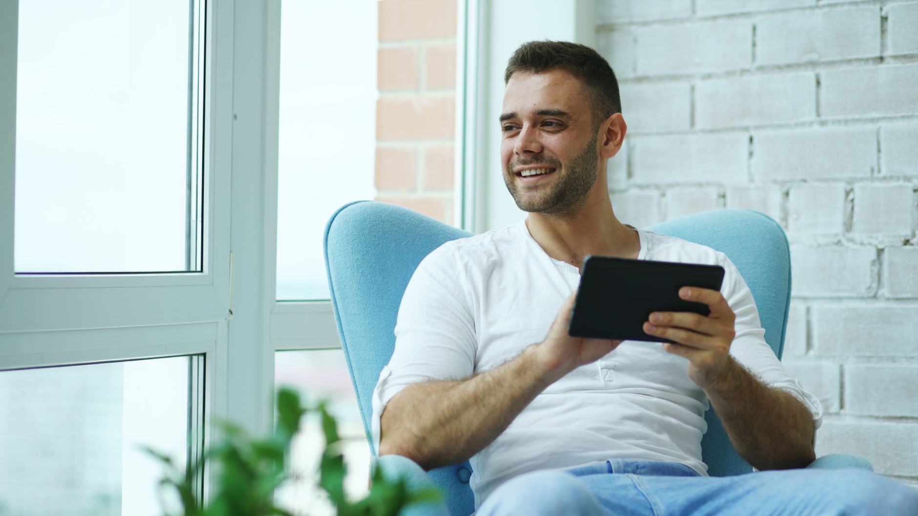 Cheerful adult man sitting by window, holding a tablet and smiling, indoors.